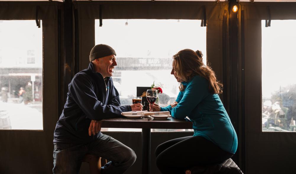 A couple sits on a covered porch and enjoy drinks while it rains outside.