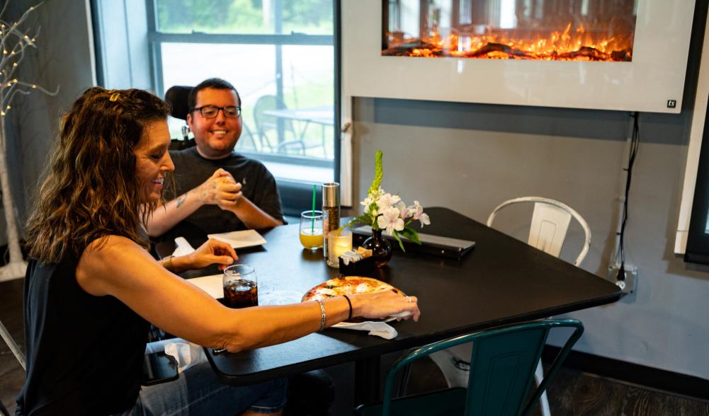 A woman and a man in a wheelchair eating pizza at a table.