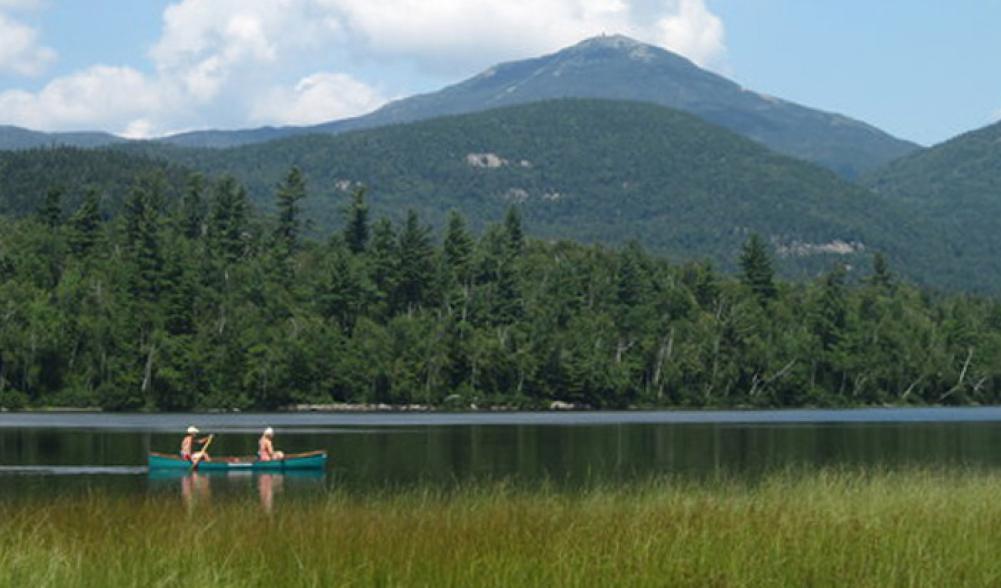 A green canoe with two people paddles on a calm lake with a large mountain in the background.