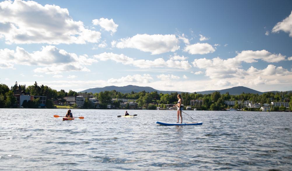 Kayakers and a woman on a SUP relax on a lake with town buildings in the background.