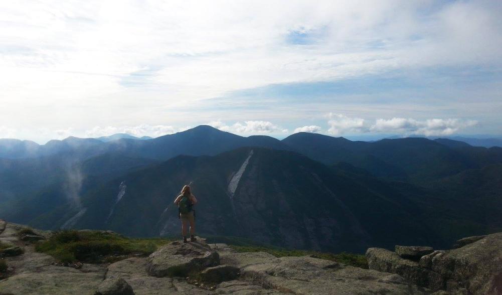 A woman with hiking backpack stands admiring the view in the High Peaks.