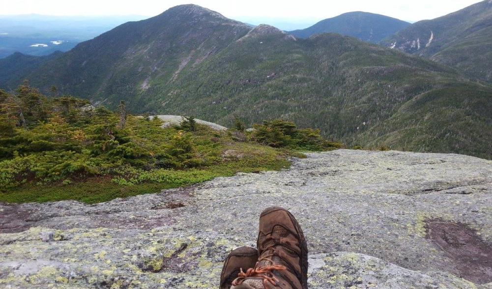 A hiker sits with legs outstretched on the rocky summit of a High Peak.