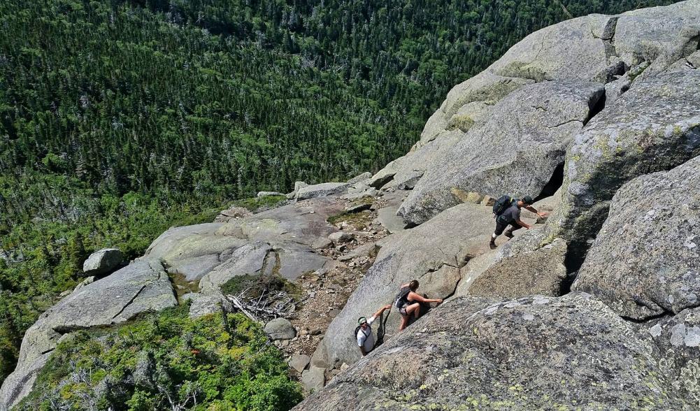 Three hikers scramble up vertical rocks on the side of a mountain.