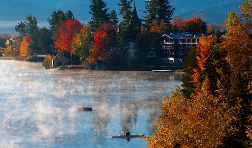 Vibrant autumn colors on the shore of a lake with fog rising over the water and a kayaker paddling.