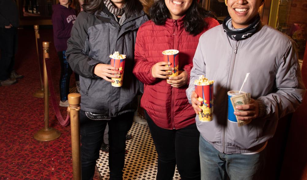 Three people stand in a movie theater lobby, smiling and holding buckets of popcorn.