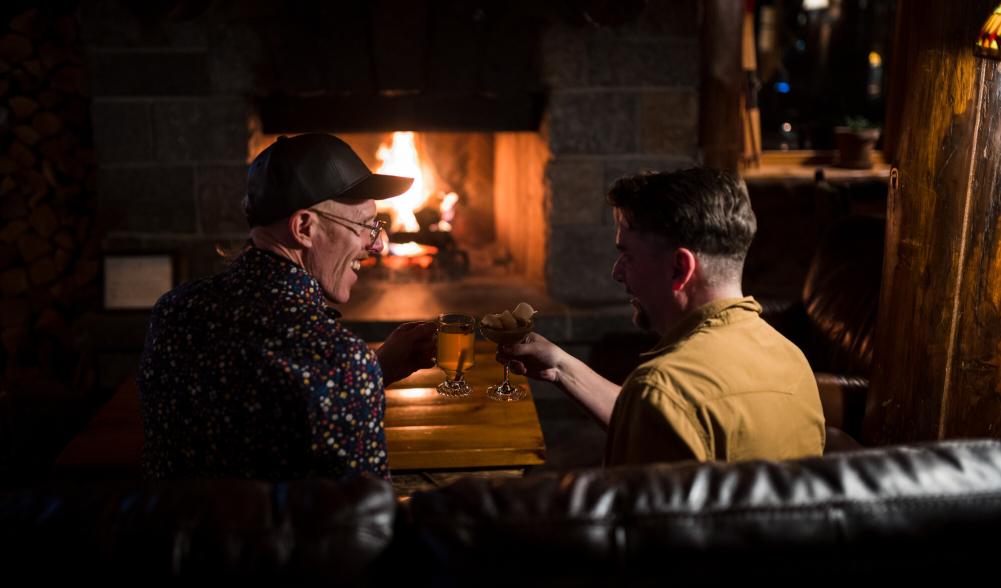 Two men cozy up by the fire with drinks in a dimly lit restaurant.