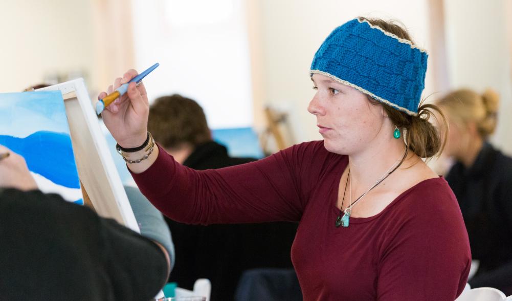 A young woman in a knit hat holds a paintbrush over a canvas.