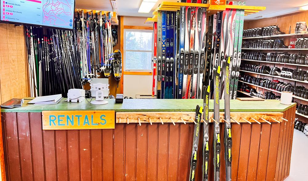 A full rental room with four pairs of skis set out for display. Boots, skis, and poles in the background for rent.