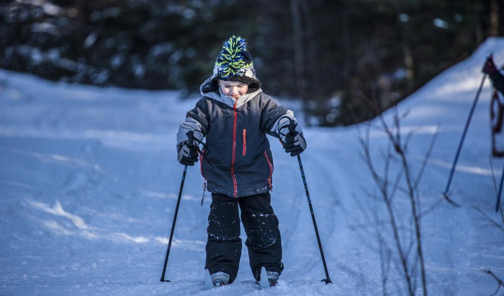 A young boy smiles and cross-country skis on a groomed trail in the woods