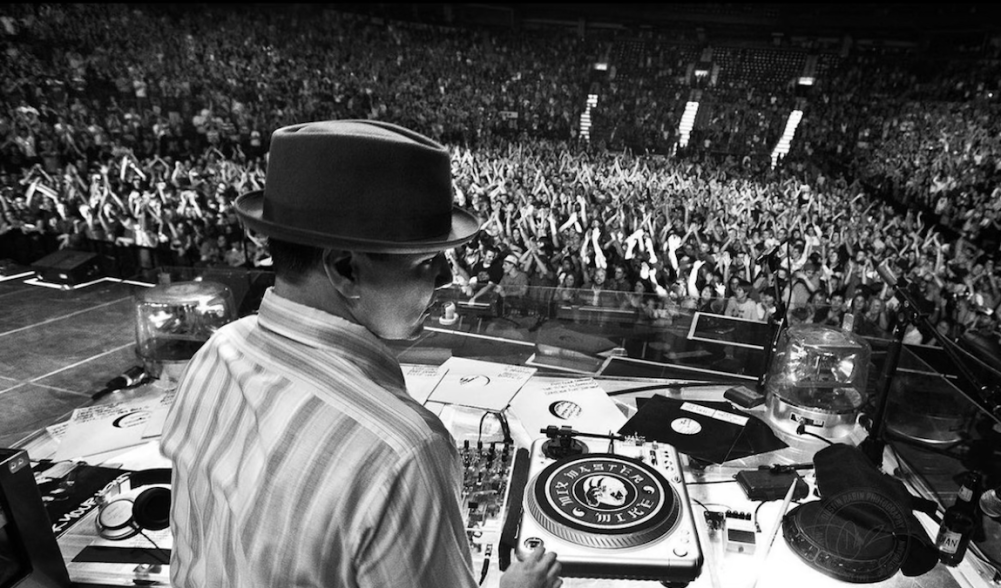 A black and white photo of a DJ wearing a fedora in front of turntables at a concert.