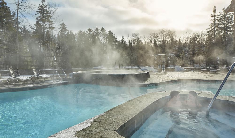 Two people sit in the water on a winter's day as steam rises from the outdoor swimming pool at the Whiteface Lodge