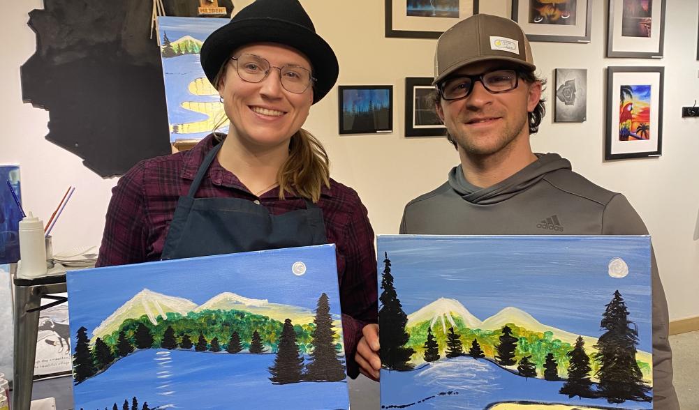 A man and a woman hold up paintings, each a version of the same image, from an art class.