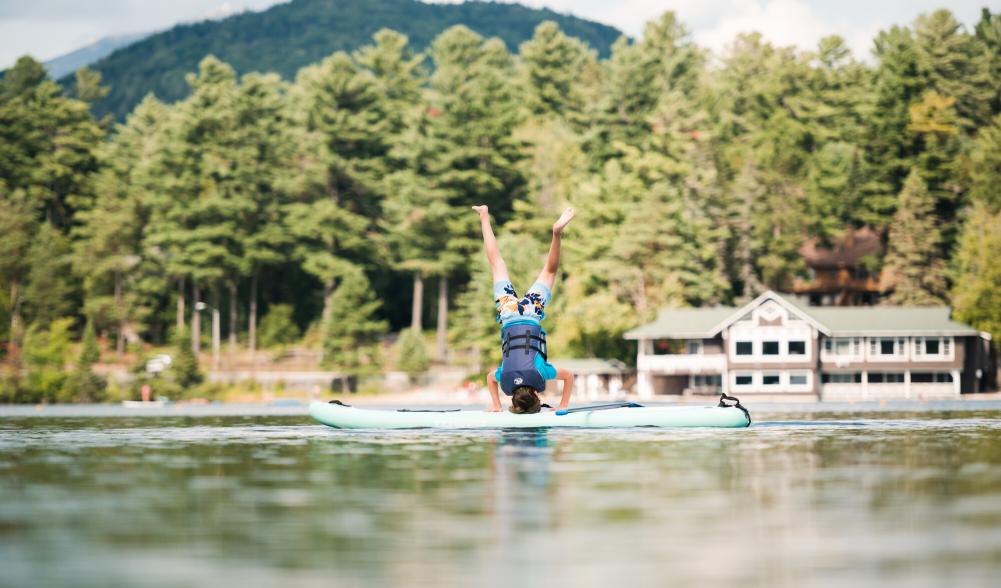 A teenager in swim clothing and a life jacket does a handstand on a stand-up paddleboard on a lake.