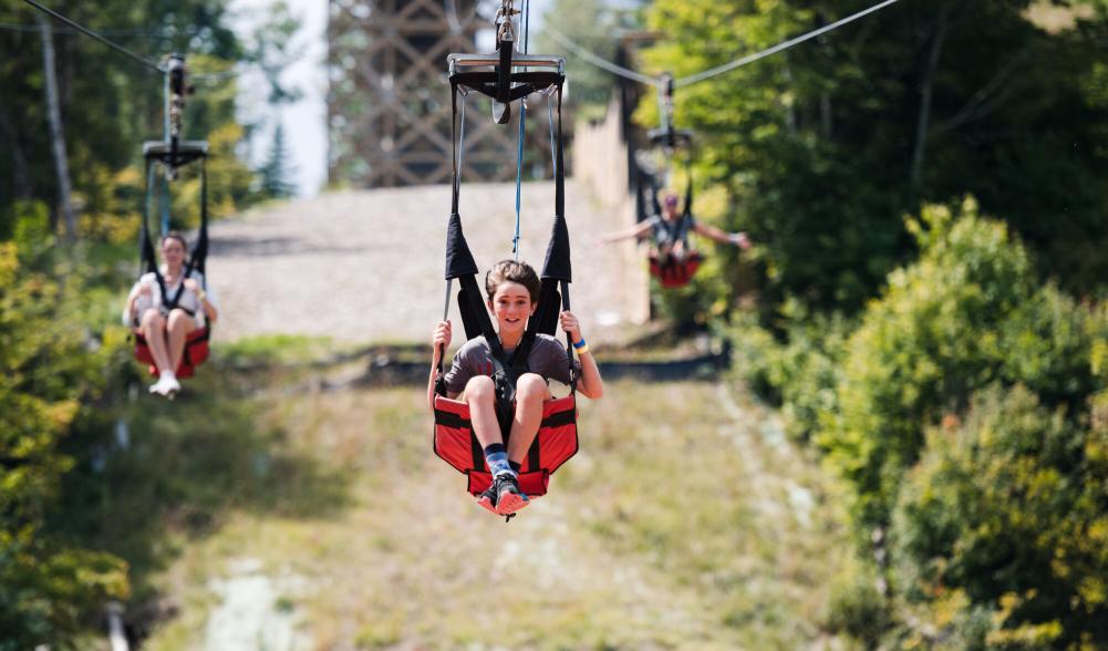 Two teenagers ride down a zipline in red and black harnesses.