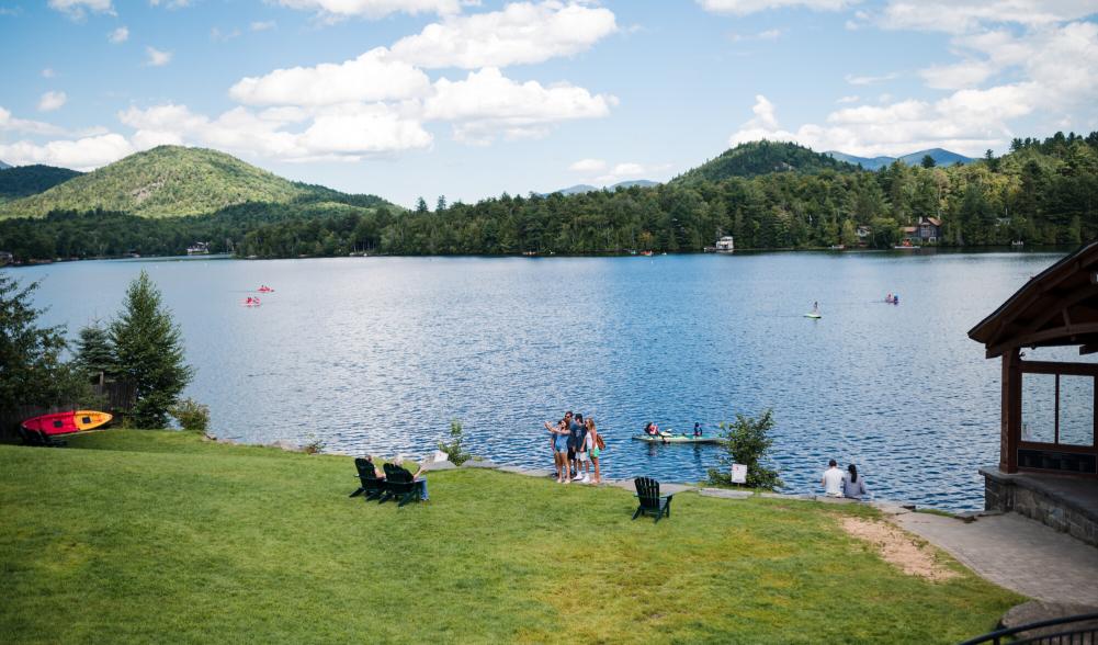 A wide angle of a sloping, grassy park in front of a bright blue lake and mountains. People dot the grass.