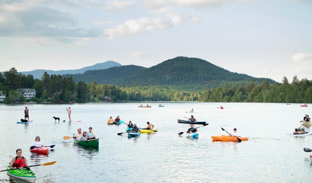 Colorful kayaks, canoes, and SUPs cluster on a sparkling blue lake with green mountains in the background.