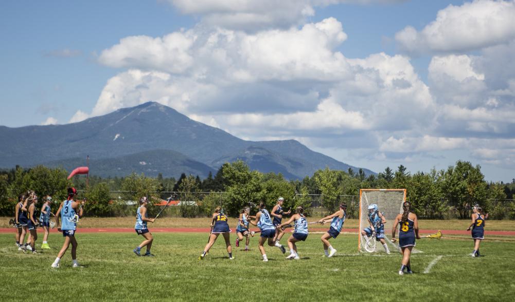 A group of athletes plays lacrosse on an outdoor field in Lake Placid, with high peaks scenery in the distance