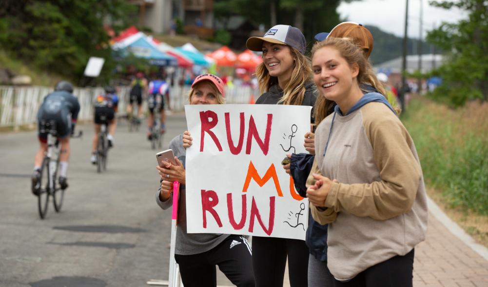 Three young women hold a sign with encouraging words for an Ironman athlete at Lake Placid Ironman