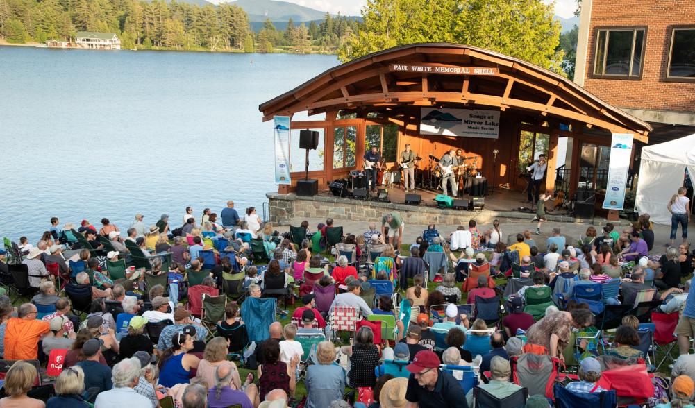 A crowd gathers near the band shell in the grass under a summer sky at Mid's Park in Lake Placid