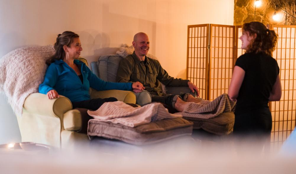 A man and woman sit next to each other on plush armchairs, with their feet up for a spa treatment from a woman with curly hair.