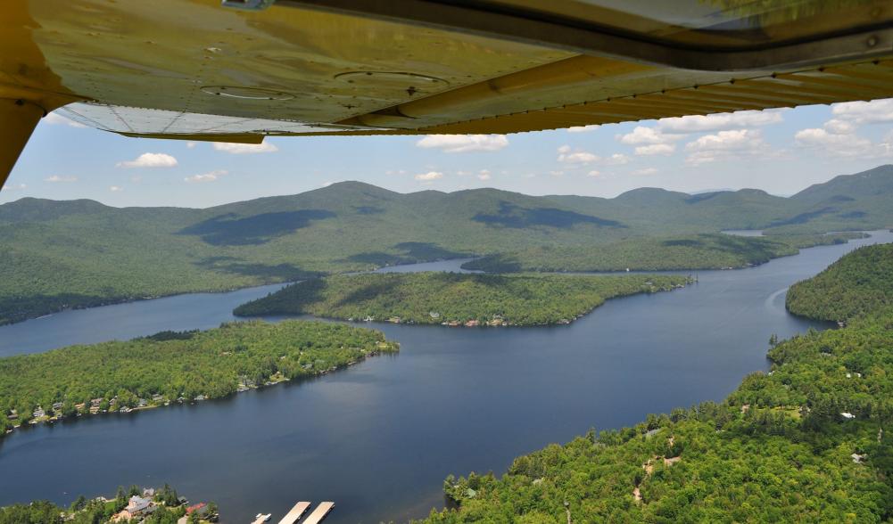 Looking down on green forests, a forested island, and blue lake from under a small yellow airplane wing.