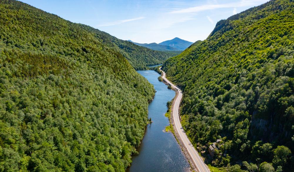 An aerial view of a long, narrow lake, surrounded by a single, narrow road and forested mountains on a sunny day.