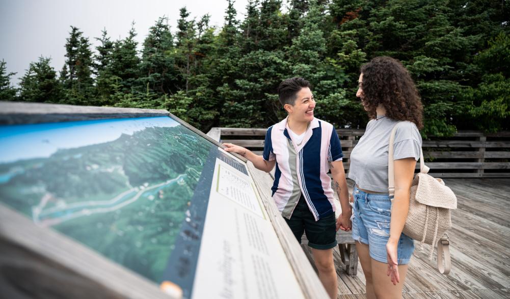 A couple stands smiling at each other in front of an interpretive display on a deck with forest in the background.