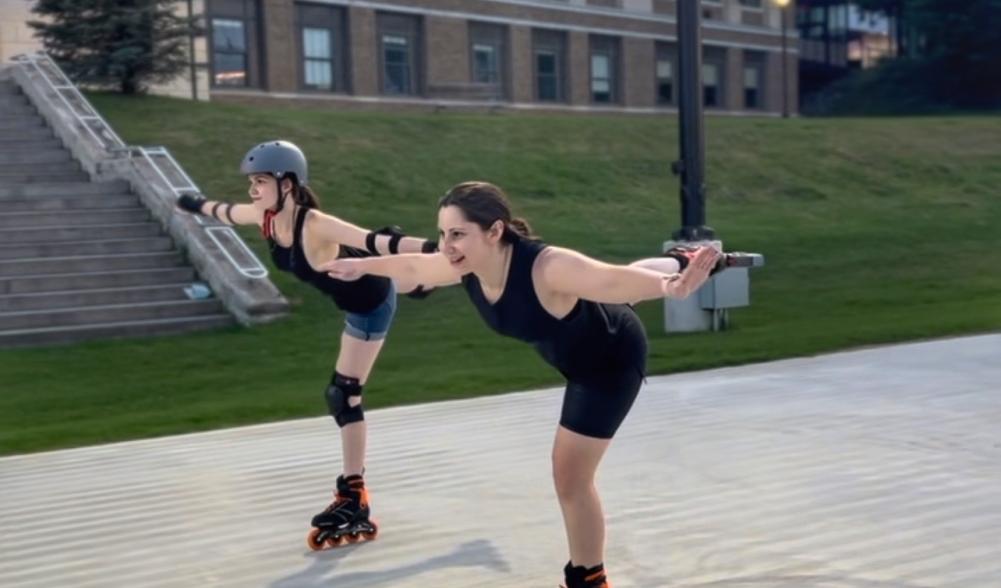 Two women inline skate side-by-side in figure skating poses, skating on one leg.