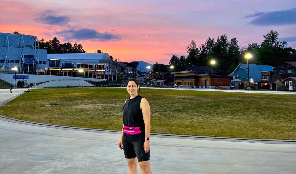 A woman stands on a skating oval wearing black and pink skating clothes and inline skates, while the sun is pink with a sunset behind her.