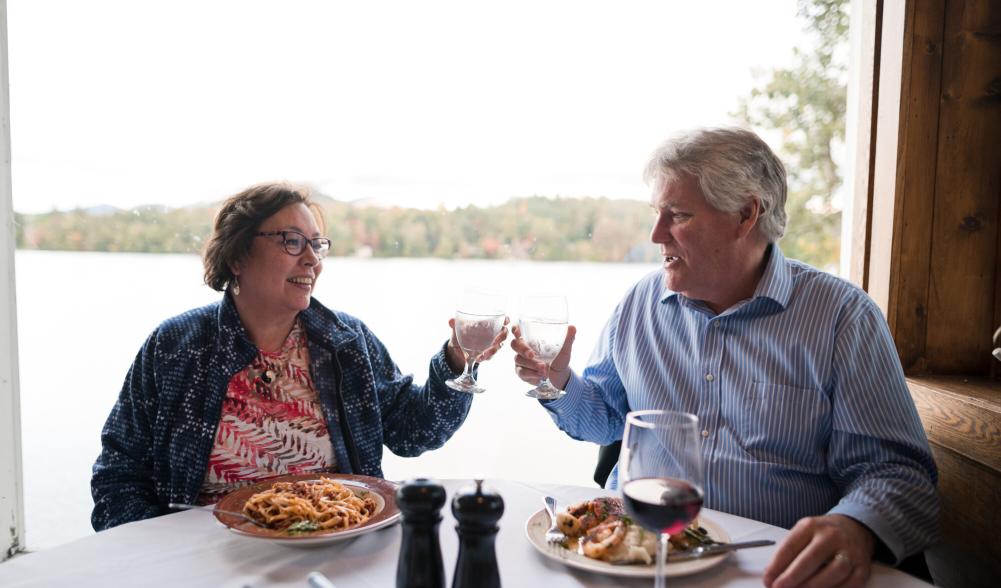 A couple (man and woman) clink drinks together at a restaurant table overlooking a lake and mountains.