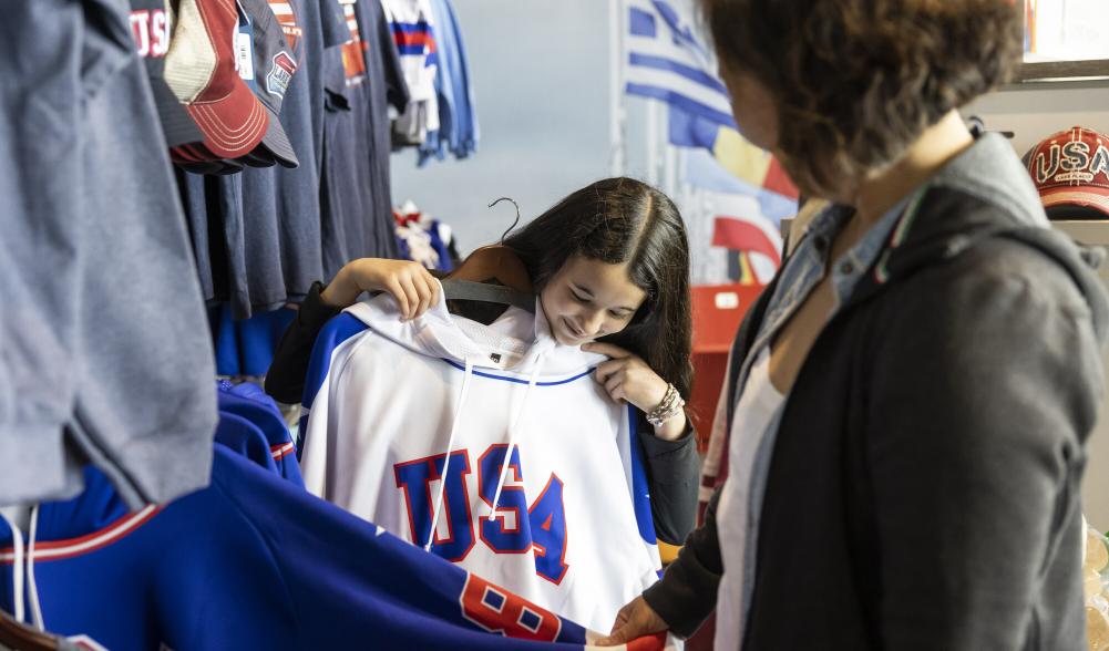 A young woman holds a USA hockey jersey up to herself in a brightly colored shop.