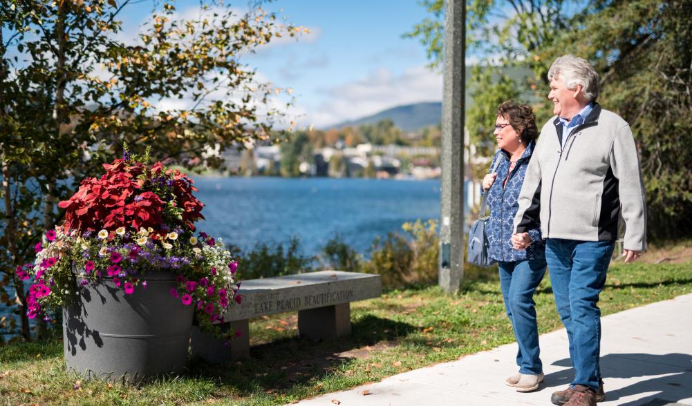 Two people walk on a sidewalk in Lake Placid village, with views of Mirror Lake in the background