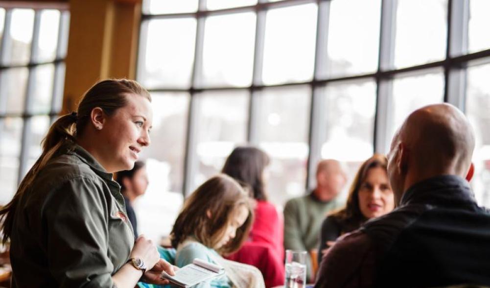 A waitress takes an order in front of a wall of windows.