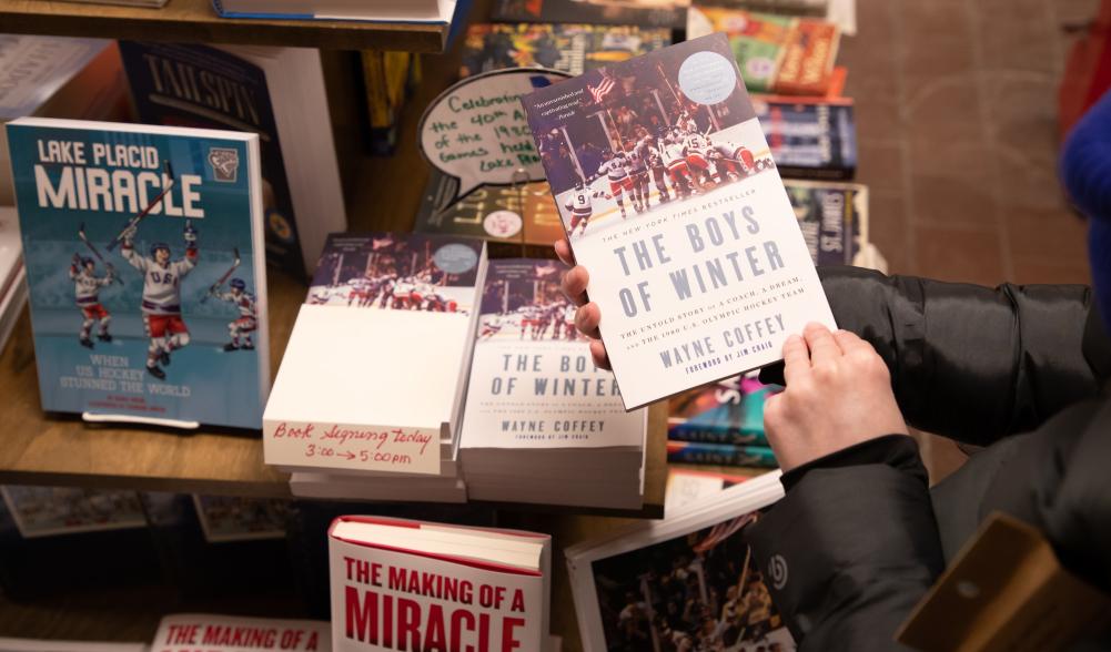 A close-up of a person's hands holding a book in a bookshop, with a book-filled table in the background.