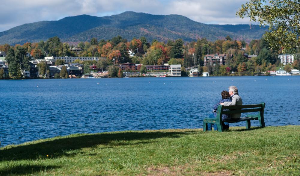 A middle-aged couple sits on a bench overlooking a lake, with a village and mountains in the background.