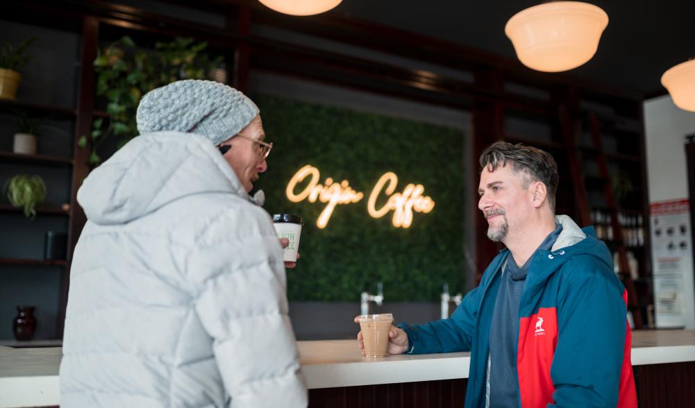 Two men share coffee together at a coffee bar with grass decor wall in the back.