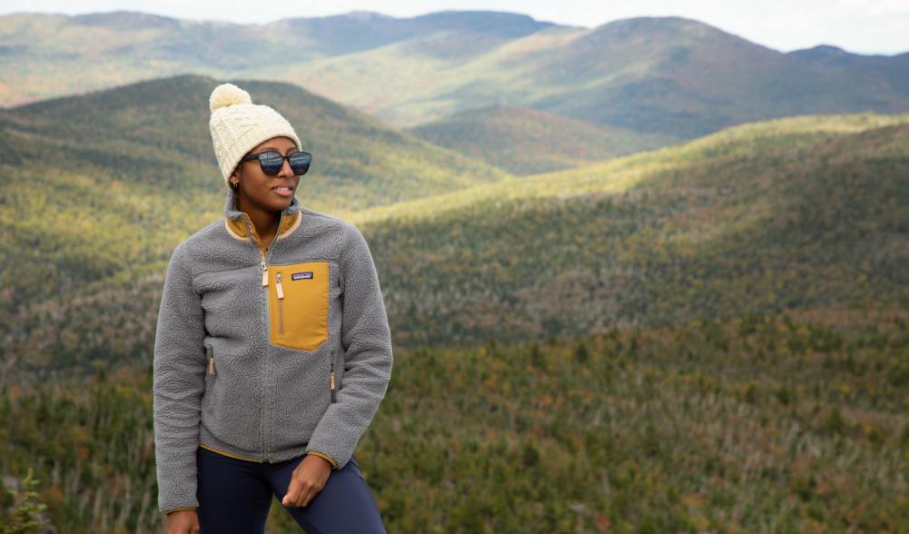 A hiker smiles facing the camera with a mountainous landscape in the background during late fall