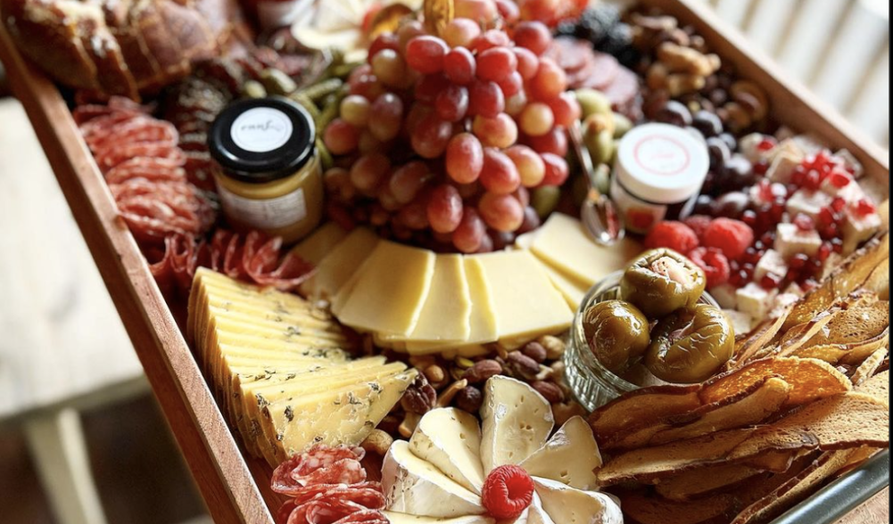 A charcuterie board with an assortment of meats, cheeses, peppers, grapes, mustard, bagel chips and more