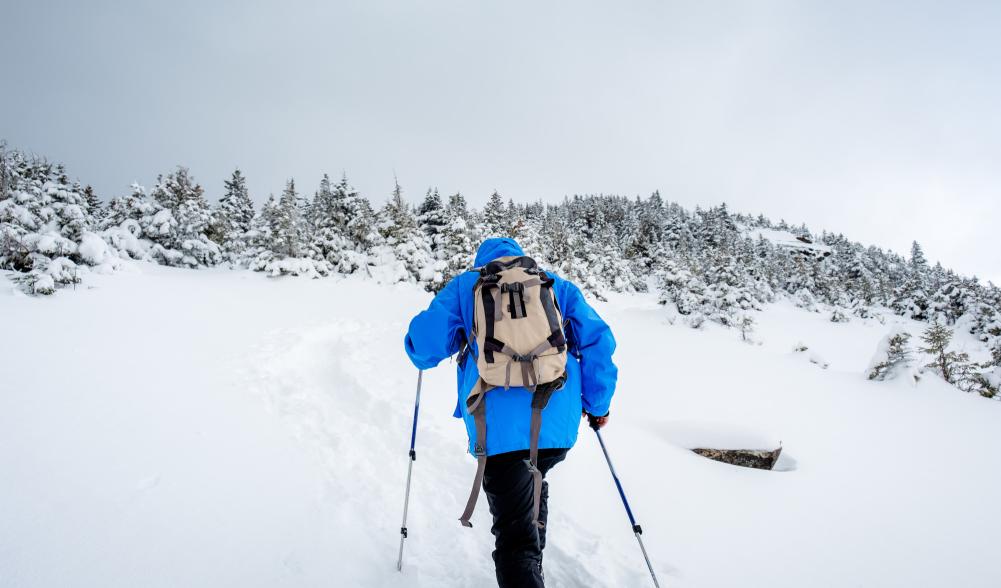 A person climbs a white snowy mountain with outdoor winter gear on and walking sticks.