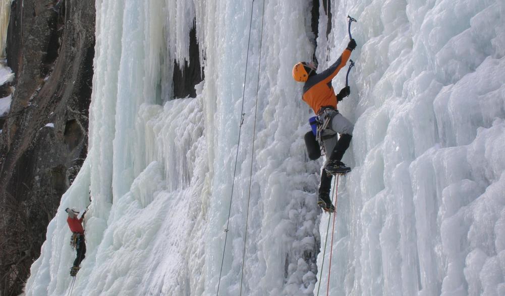 Two people scale an icy wall on a mountain with pick axes and harnesses.