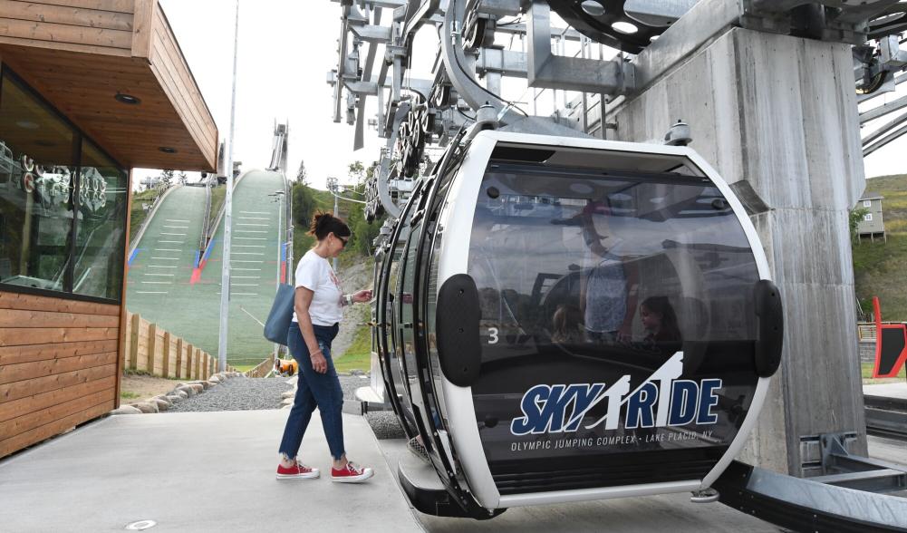 A woman steps into a gondola in front of green ski jumps.