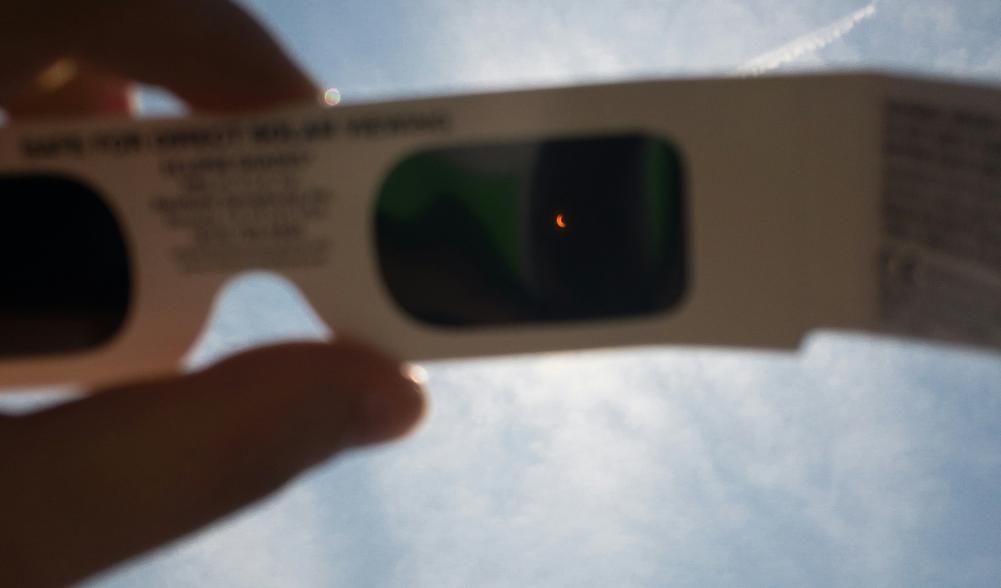 A partial solar eclipse is visible through eclipse glasses held up to a sunny sky.