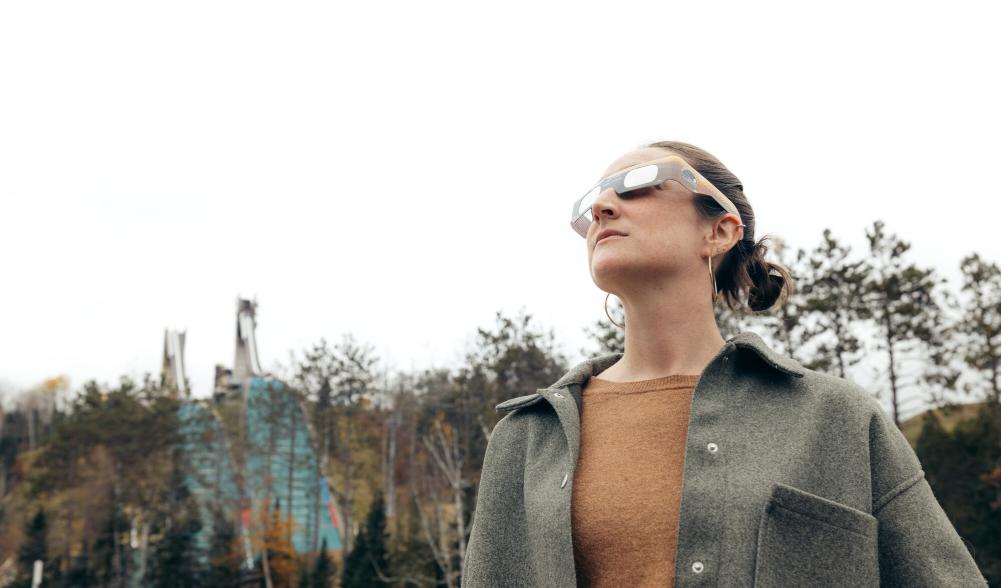 A woman wearing eclipse glasses gazes up at the sky with ski jumps in the background.