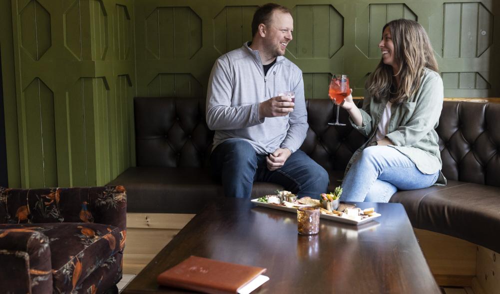 Couple sits on bench seat with cocktails and food on table