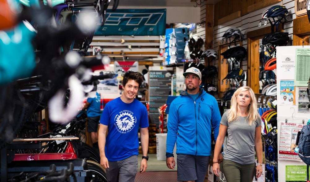 A bike shop employee leads a man and a woman to a display of bikes in a crowded shop.