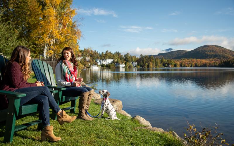 Two women and a dog sit in Adirondack chairs next to a calm lake with fall foliage around.