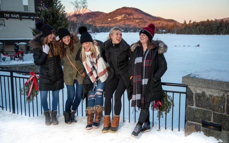 Five women in winter clothing laugh in front of a snowy lake and sunset-lit mountains beyond.