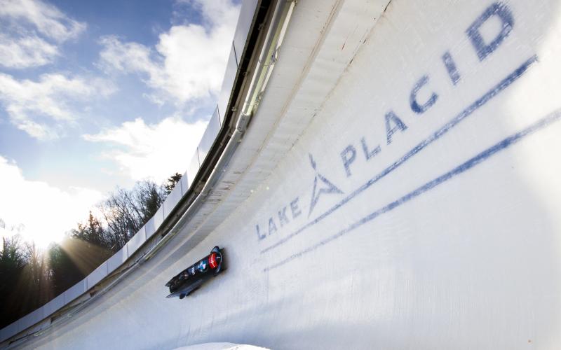 A team takes a large banked curve on the Lake Placid Olympic bobsled track with white clouds and blue sky in the background