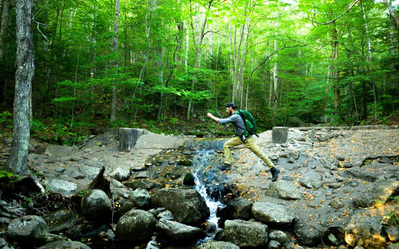 A person in yellow pants hops across a small stream while hiking on Haystack Mountain