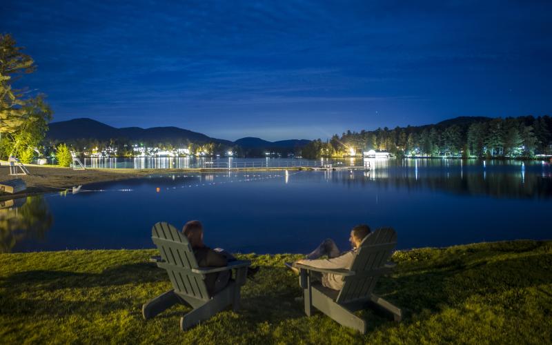 A nighttime view of the lake with people sitting on Adirondack chairs.
